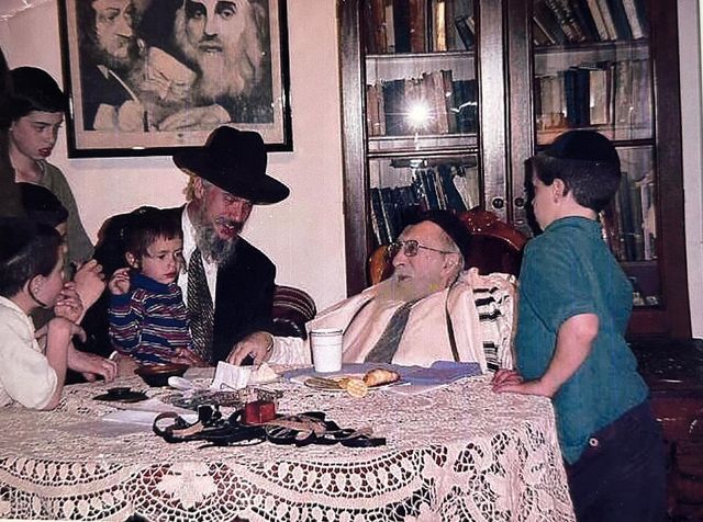 Rabbi Yisroel Meir Vogel at a table with Rav Pinchas Scheinberg, in a tallis, and children.